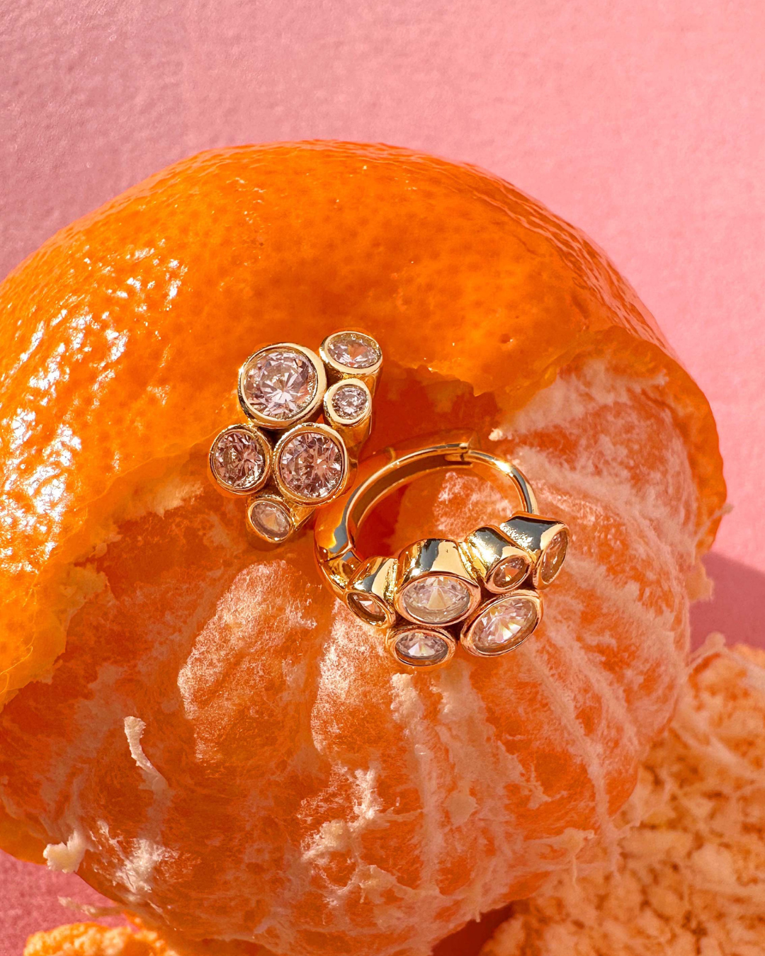 Romyli Bubble Cluster Huggies in 18k gold vermeil with bezel-set CZ cluster, styled on citrus fruit against a pink background