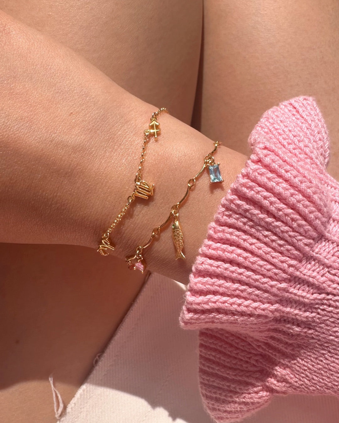 Personalised Gold bracelet with charms on a wrist and Petite Zodiac Bracelet, wearing a pink sweater.