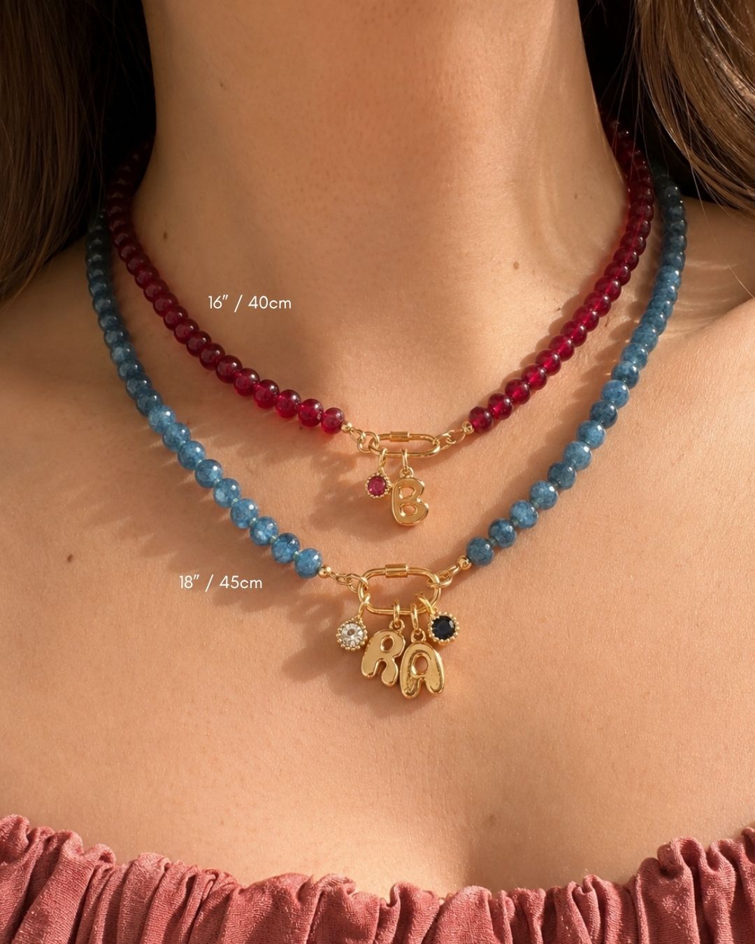 A close-up of a model's neck and upper chest wearing two gemstone beaded necklaces layered over a dusty rose top. One necklace is made of dark red jade beads with a golden charm holder clasp and bubble initial and birthstonecharm, while the other is made of blue jade beads with a gold 'BA' initial pendant and birthstone pendants.