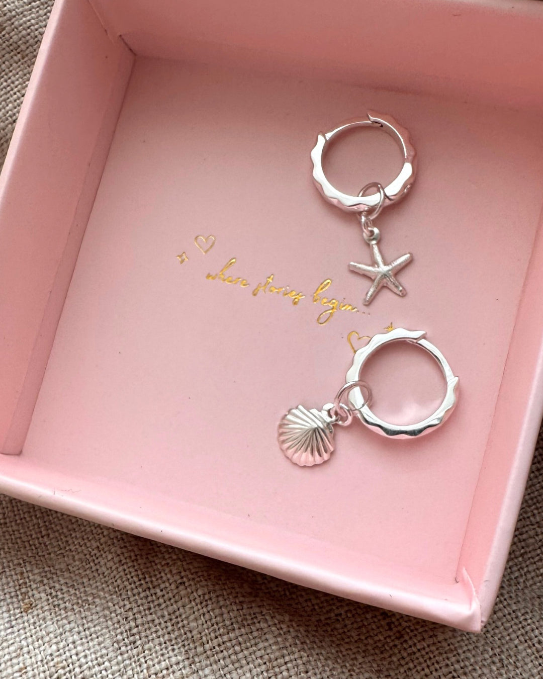 Sterling silver huggie hoop earrings with a hammered finish and starfish and seashell charms, displayed in a ROMYLI gift box