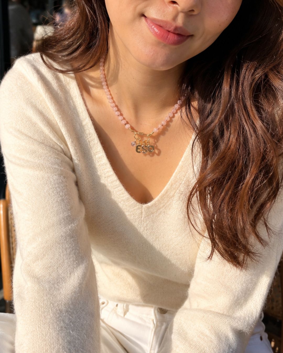 A close-up lifestyle shot of a woman wearing a pink Jade beaded necklace with a gold initial 'E' pendant and birthstone pendant, paired with a cream-colored knit sweater.