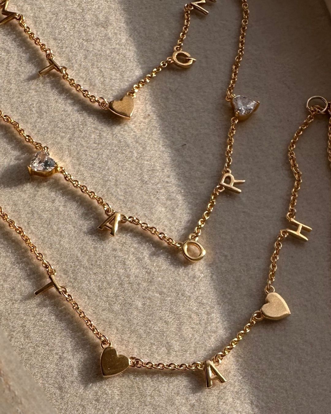 Gold initial bracelets with heart and letter charms on a neutral surface
