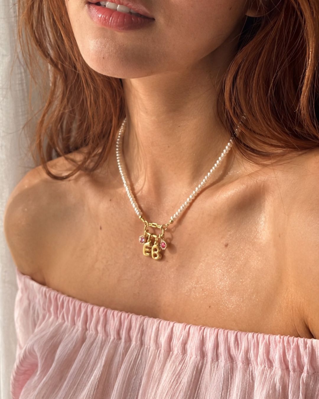 Woman wearing a pearl charm holder necklace with gold charm pendants and birthstone pendant and 'EB' letters, pink top.