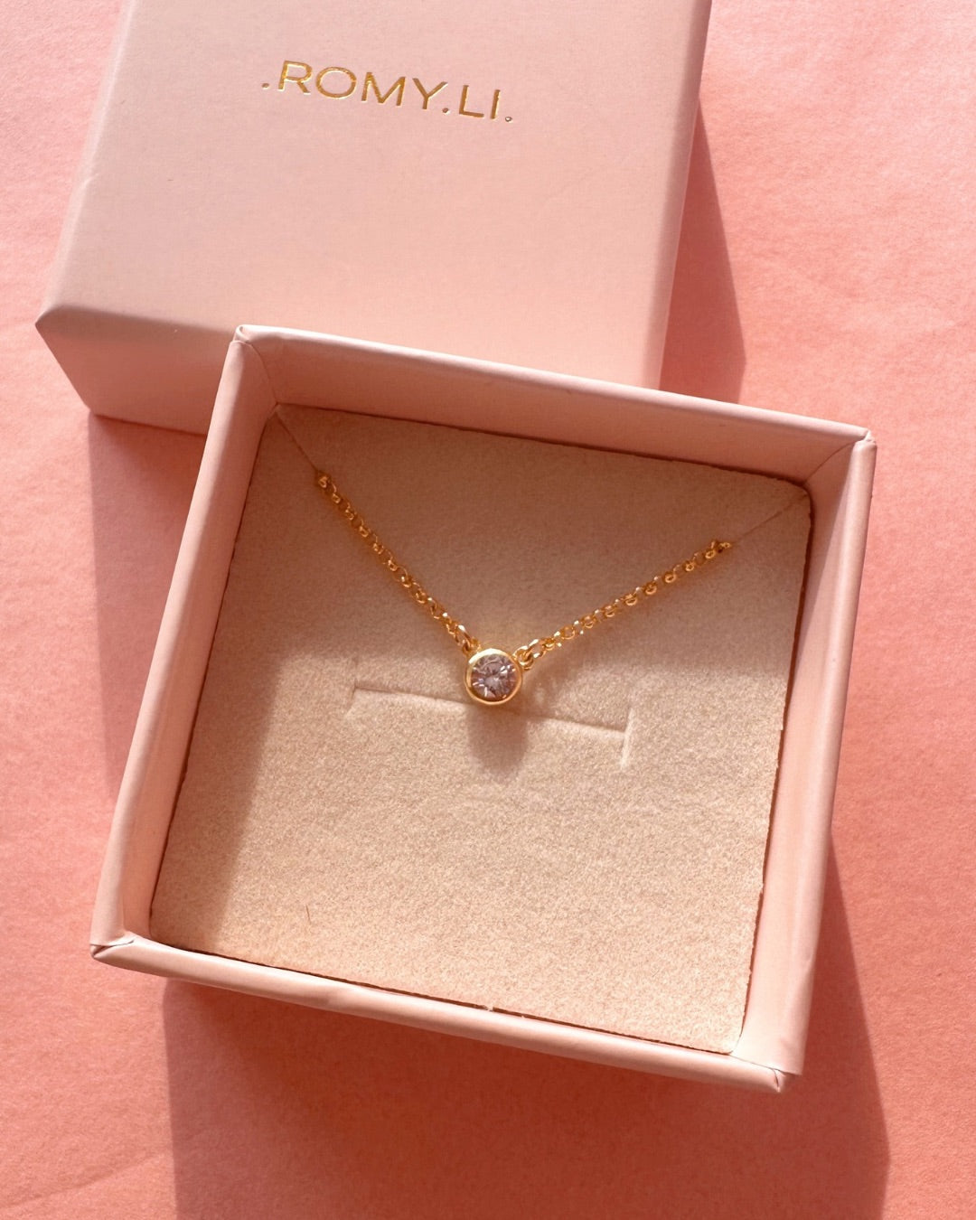 Gold necklace with a diamond pendant in a pink 'ROMY.LI.' box on a pink background