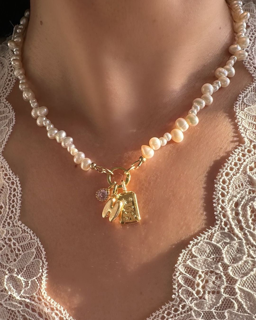 Pearl necklace with a gold bubble initial and birthstone pendant charm on a person wearing a lace garment.