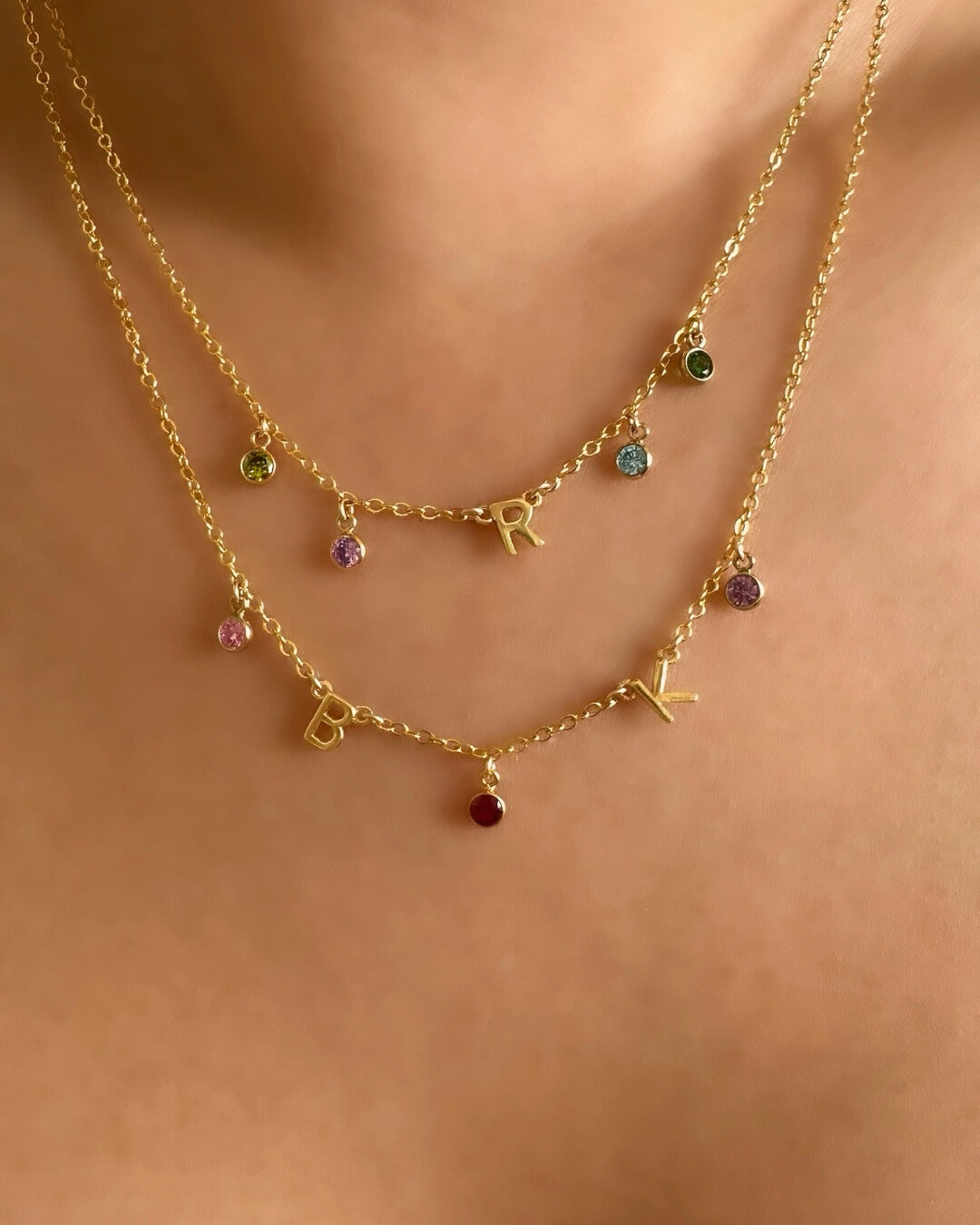 A dainty gold initial birthstone necklace on a model, featuring personalised letter and birthstone charms, elegantly displayed on a fine gold chain. Perfect for everyday wear and meaningful gifting.