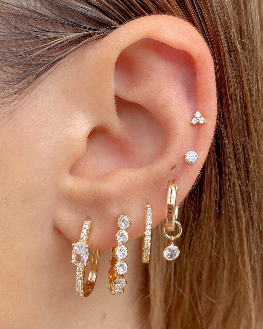 Gold cubic zirconia square pave hoop earrings stacked with gold hoop earrings and diamond stud earrings and Flatback earrings on a model