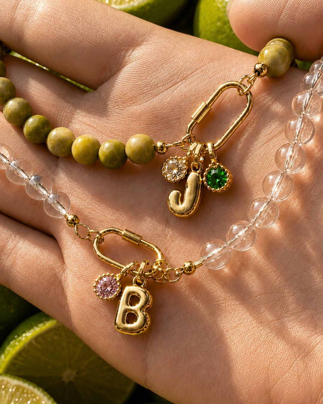 A close-up of a hand wearing a gold-tone beaded necklace with round crystal quartz beads, olive-green Jasper beads, and two letter charms: a gold 'J' charm with a November birthstone (citrine) and a green May birthstone, and a gold 'B' charm with a pink October birthstone (tourmaline). Lime slices are visible in the background.
