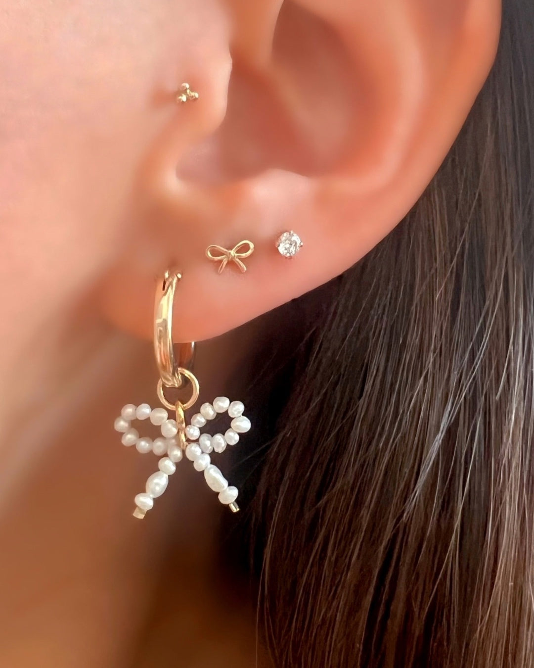Gold Freshwater Pearl Bow Hoop Earrings stacked with solid gold bow stud earrings, solitaire cubic zirconia crystal stud earrings and solid gold trinity stud Flatback earrings on a model
