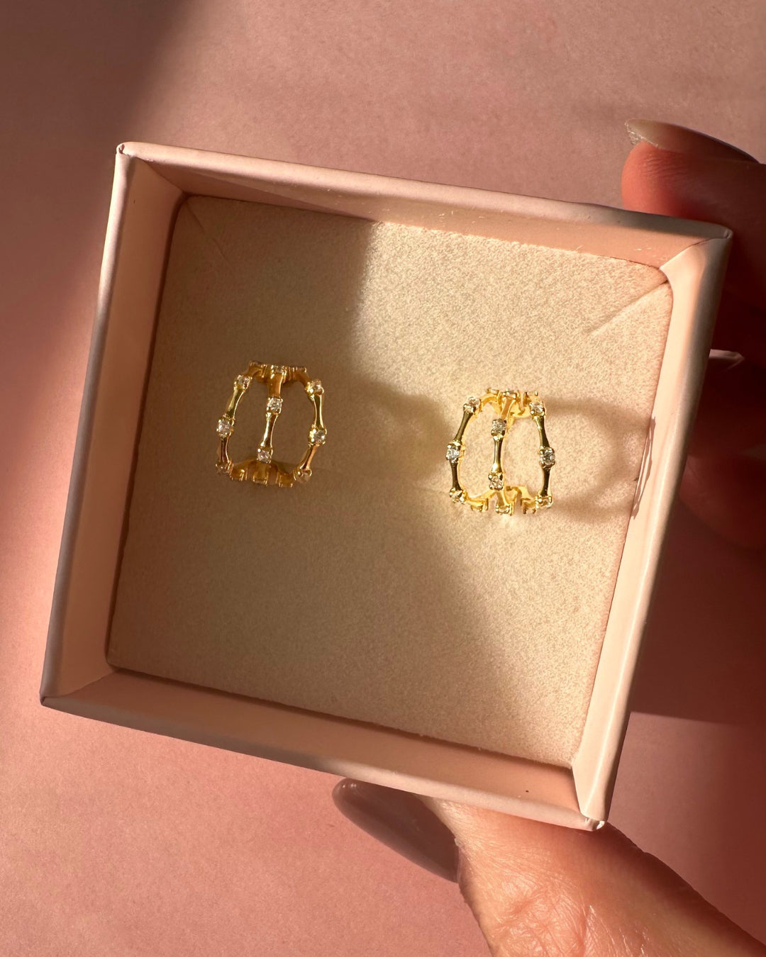 Gold trellis huggie hoop earrings in a Romyli gift box held by a hand on a pink background