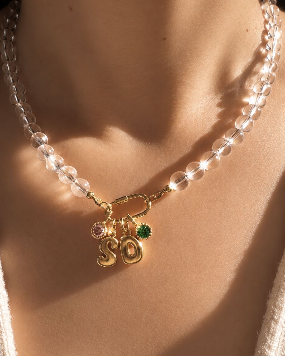 A close-up of a person's neck and chest wearing a crystal quartz beaded necklace with a gold-tone 'SO' bubble initial charm pendants and small pink and green birthstone pendants.