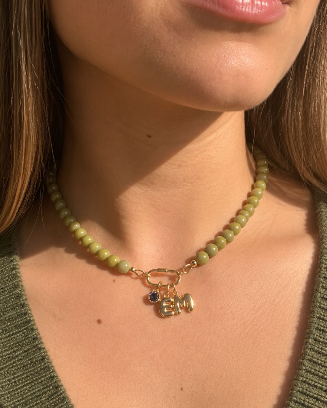 A close-up of a woman wearing a green Jasper beaded necklace, a gold carabiner clasp, and small pendants including a blue December birthstone and a gold bubble initial pendants. 