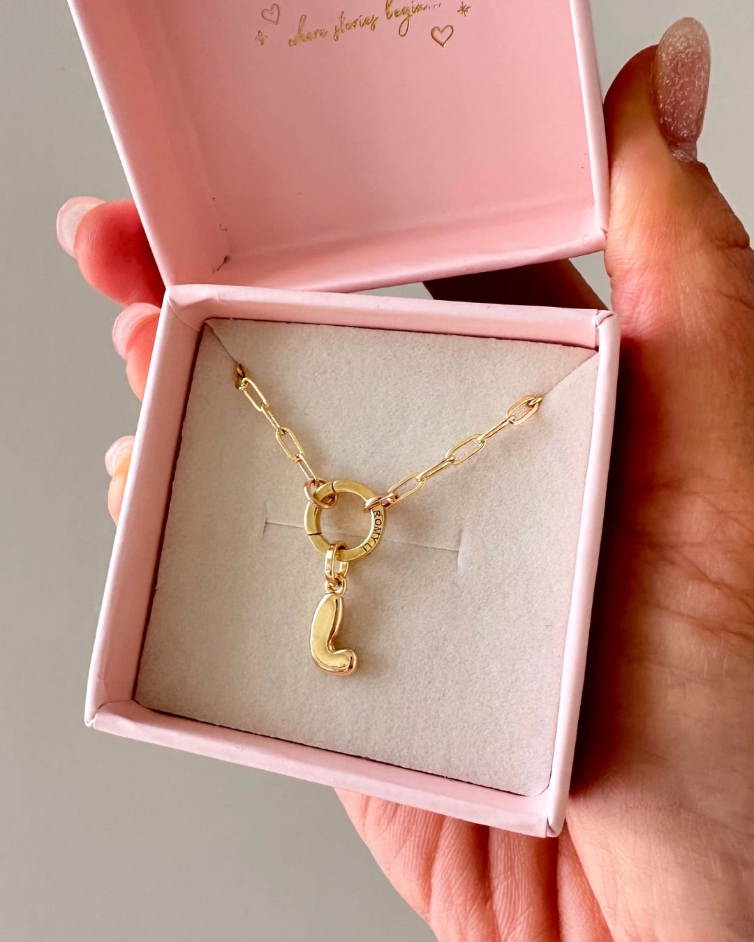 Hand holding gold Paperclip Charm Holder Necklace with bubble initial L pendant in a pink Romyli gift box against a grey background 