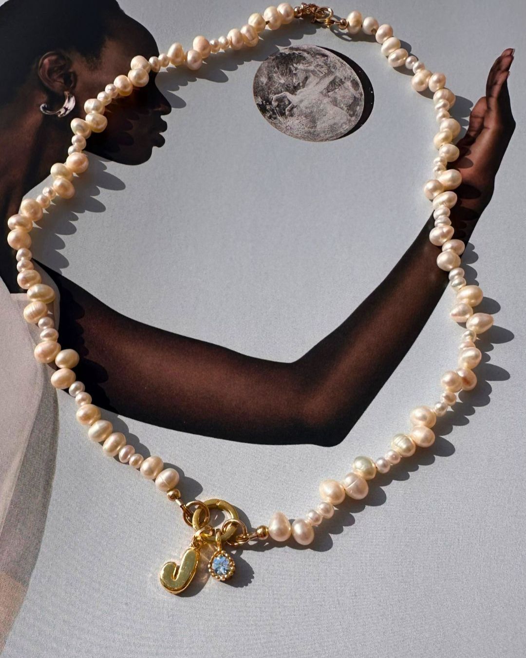 Pearl necklace with a gold bubble initial and birthstone charms held by a person against a neutral background