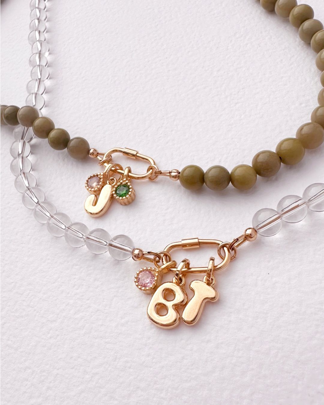 A close-up of a layered beaded necklace with olive green Jasper round beads, a clear crystal quartz bead chain, and gold charms including bubble letters 'B' and 'T', a small green birthstone charm, and a pink birthstone charm on a textured white background.
