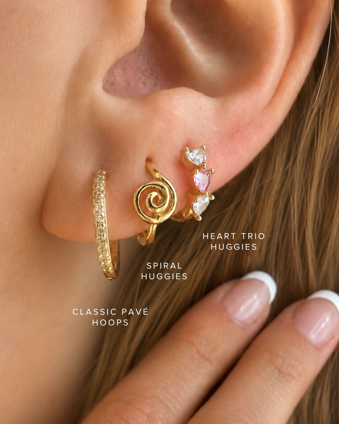 Close-up of a person's ear wearing multiple gold earrings: a classic pave hoop, a spiral huggie, and a heart trio stud with clear and pink stones. White text labels identify each earring.