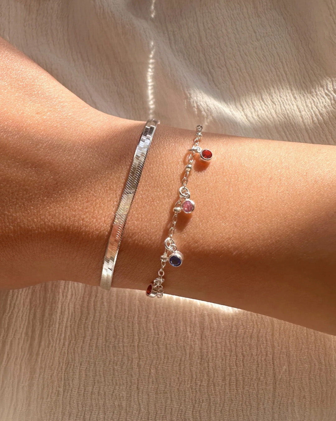 Silver dainty birthstone bracelet with up to 9 personalised cubic zirconia charms on a delicate sterling silver chain on a model. Stacked with the Silver Serpentine Bracelet