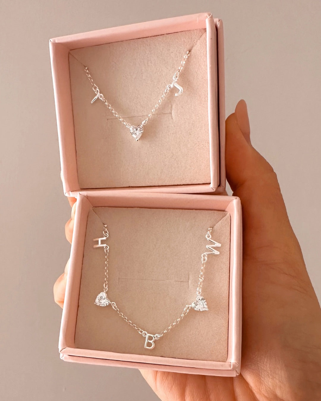 Silver necklaces with heart and letter pendants in a pink jewelry box held by a hand.