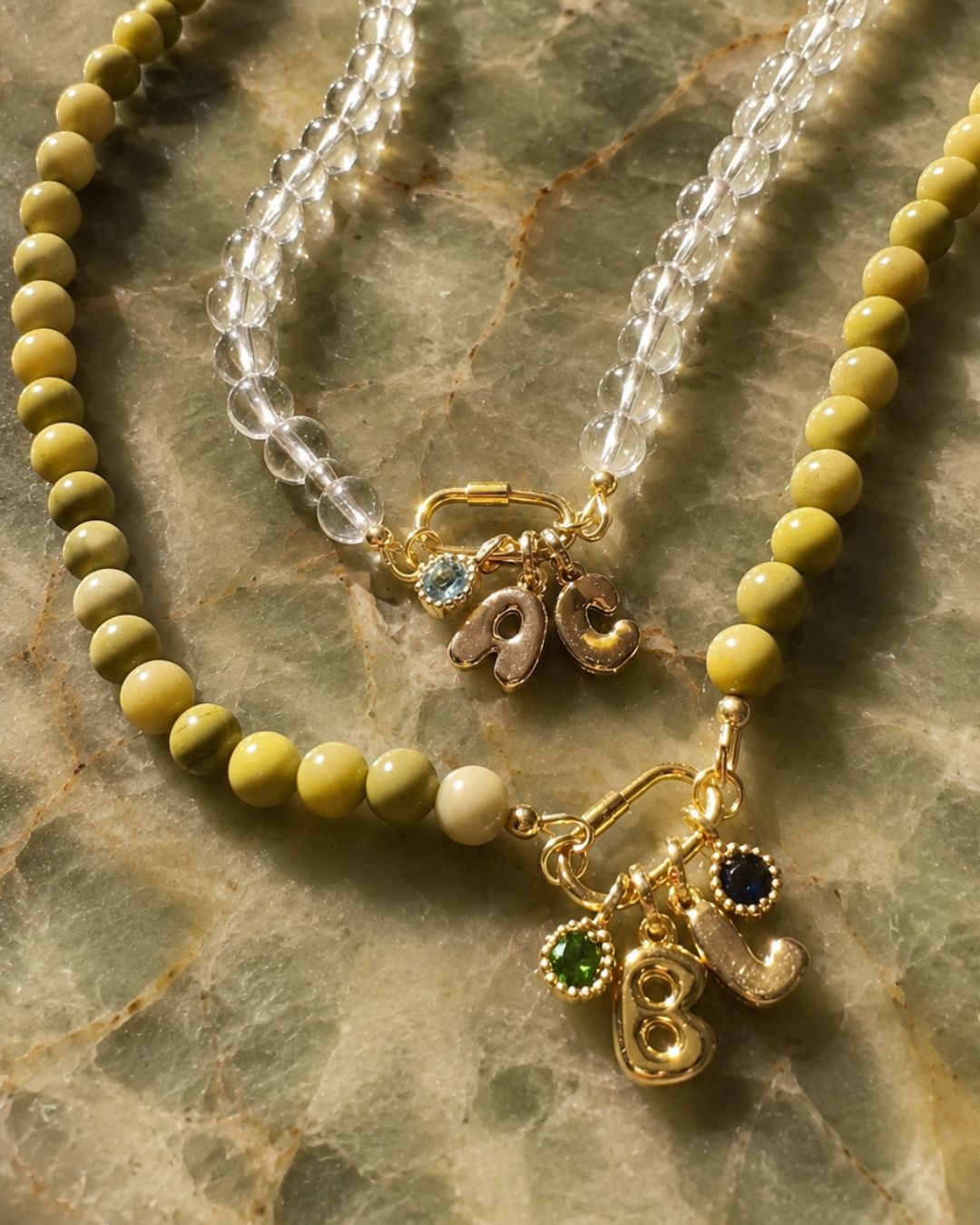Two beaded necklaces displayed on a green marble surface. One necklace features olive green round Jasper beads with gold-toned letter charms (B and L) and the other features crystal quartz crystal beads with gold-toned letter pendants and birthstone pendants. 