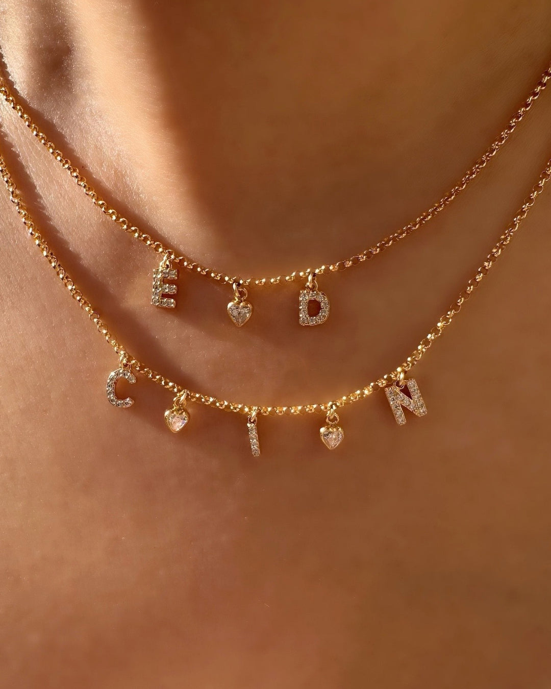 Model wearing layered gold pave initial necklaces with personalised letters and tiny bezel heart charms, dainty gold jewellery for everyday wear