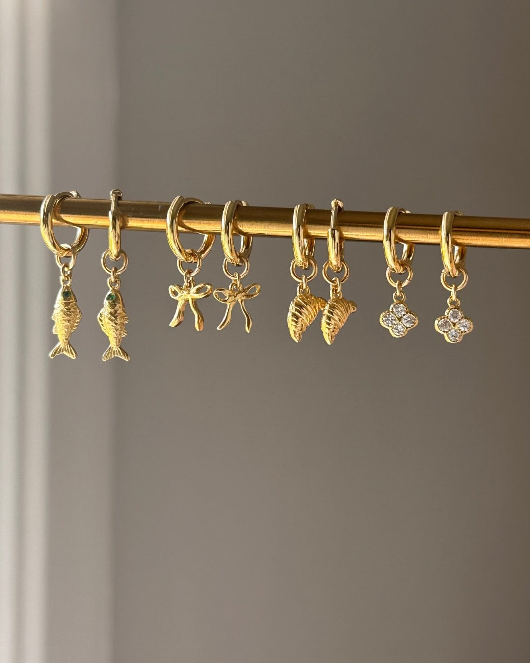 Set of gold charm hoop earrings with bow, sardine, mini clover and croissant charms on a rod against a neutral background