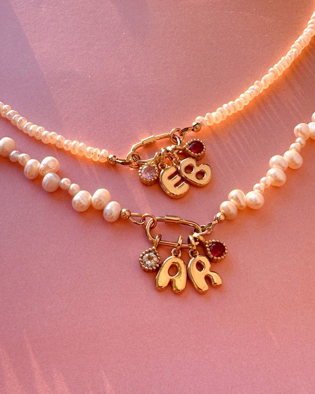 Two pearl charm holder necklaces with gold pendanf charms on a pink fabric background