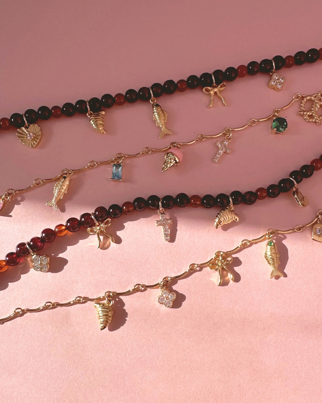 Garnet charm necklaces and bracelets with gold curve bar charm necklace and various charms on a pink background