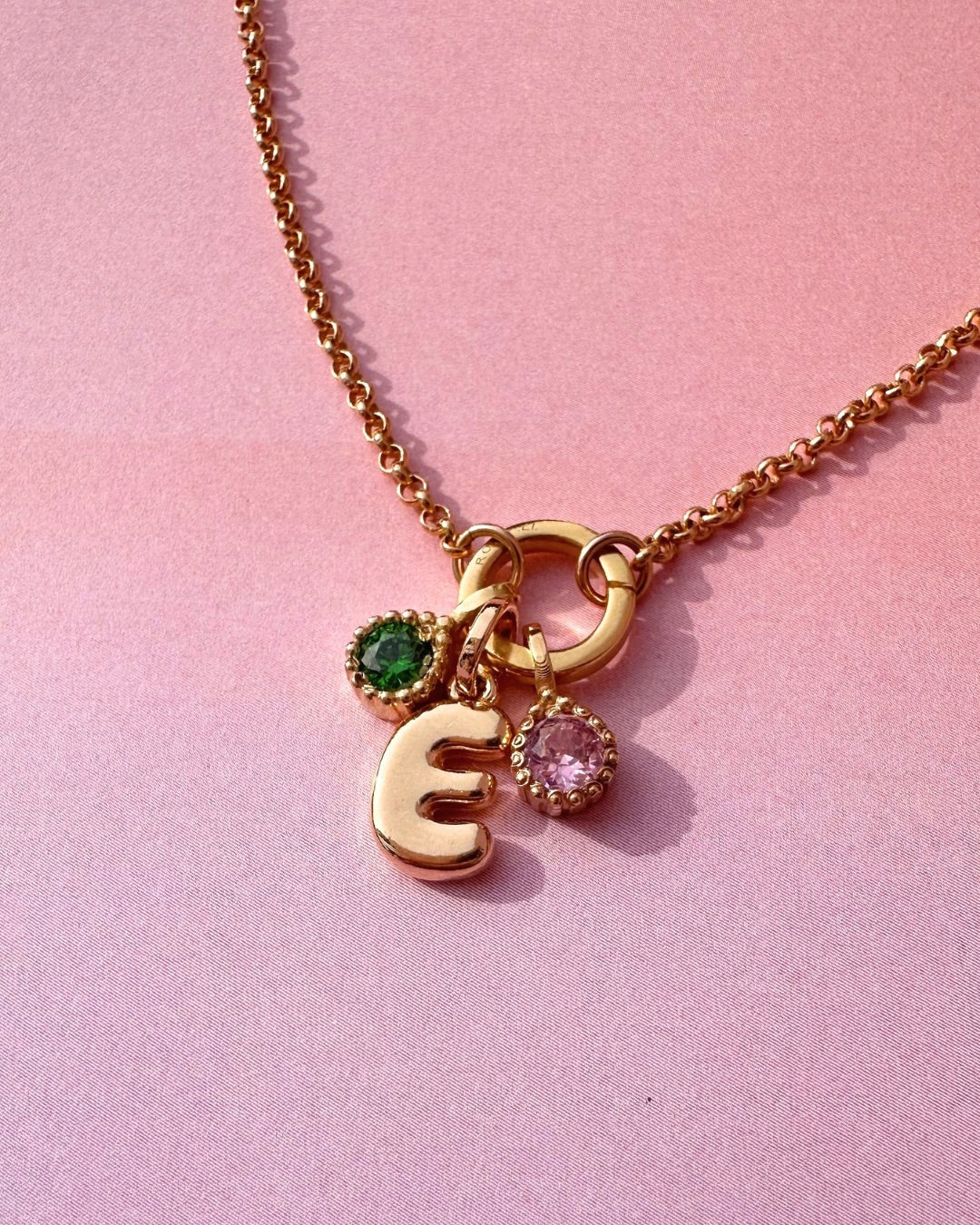 Gold necklace with bubble letter 'E' charm and two birthstone pendants on a pink background