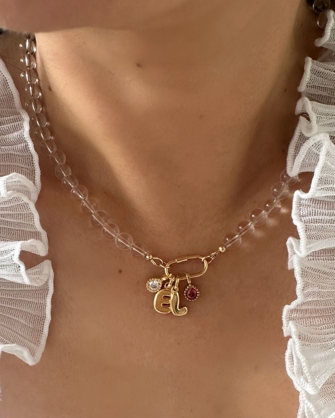 A close-up of a person's neck and collarbone wearing a clear crystal quartz bead choker necklace with a gold clasp and dangling charms, including a gold initial 'E' and small birthstone pendants, paired with a white ruffled top.