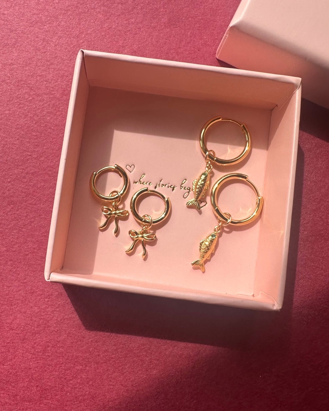 Gold charm hoop earrings with bow and sardine charms in a pink box on a red background